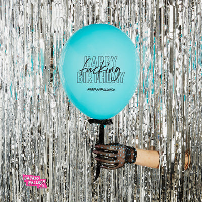 Woman holding fun party balloon with sayings Happy Fucking Birthday in front of a silver disco background 